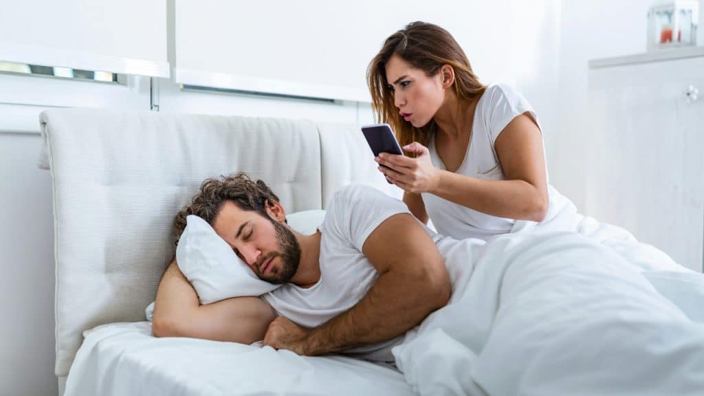 A woman holds a phone over a man sleeping in bed, looking upset at him.