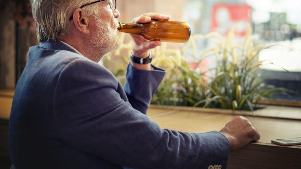 A bearded man in a blazer sits at a table drinking from a bottle.