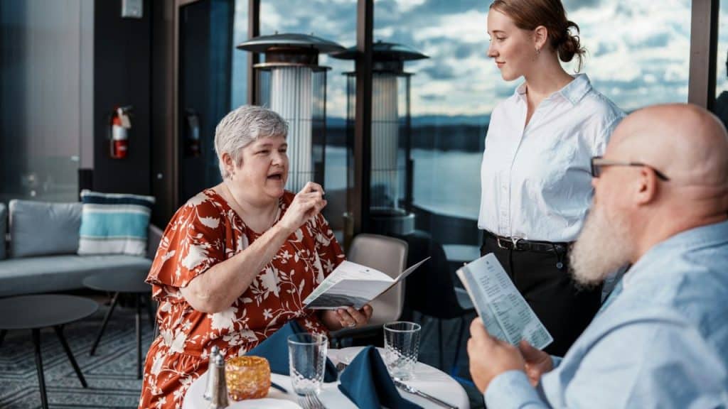 A woman holds a menu while speaking to a server at a restaurant table.