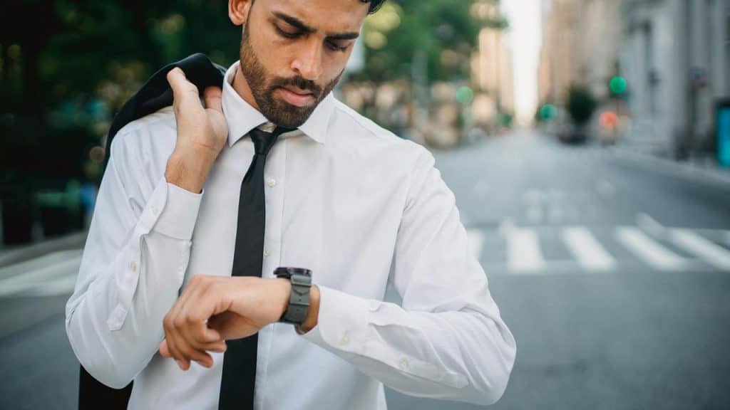 A man in a shirt and tie checking his watch on a city street.