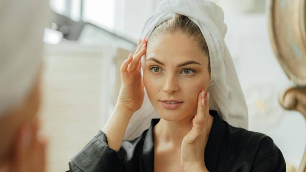 A woman with a towel on her head touching her face while looking in a mirror.
