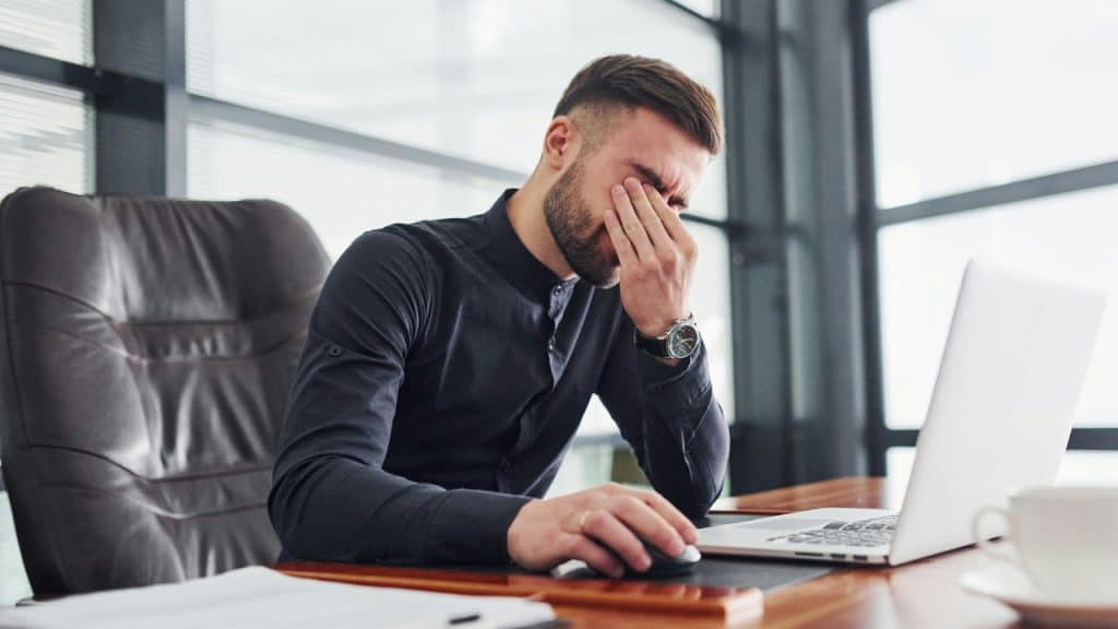 A man in a black shirt sits at a desk, rubbing his eyes near a laptop.