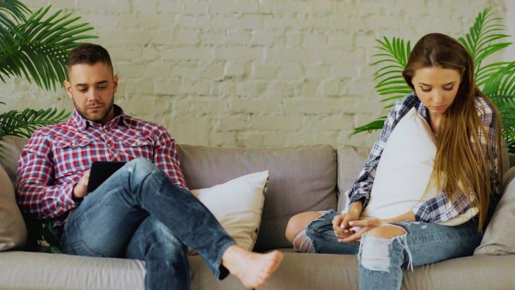A man and woman sit on a sofa, looking down away from each other.