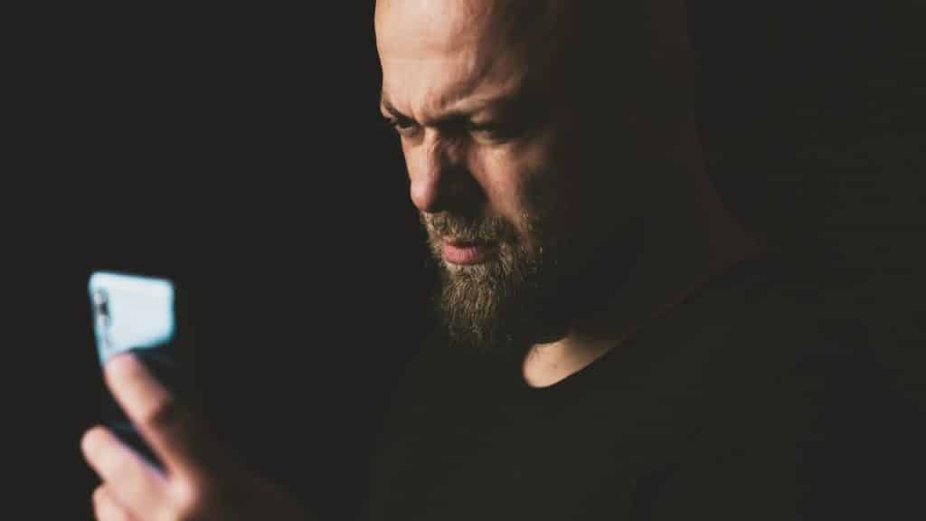 A bearded man looks intensely at the bright screen of his dark smartphone.