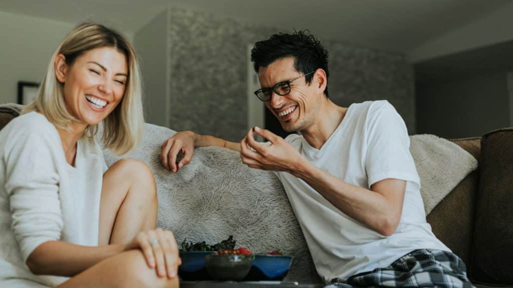 A man and woman sit on a gray sofa laughing while eating snacks from bowls.