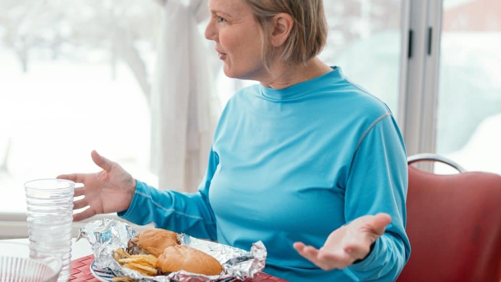 A senior woman sitting at a table with a partially eaten sandwich and a glass of water, gesturing with her hands as if explaining something or having a conversation indoors near a window.
