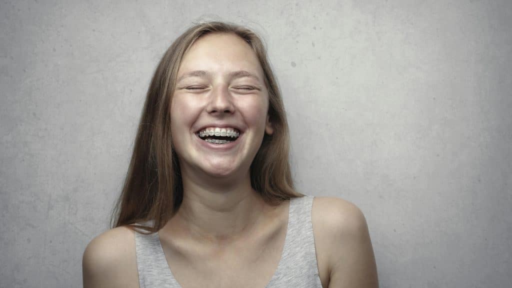 A young woman with braces laughs with her eyes closed against a plain, textured gray wall.