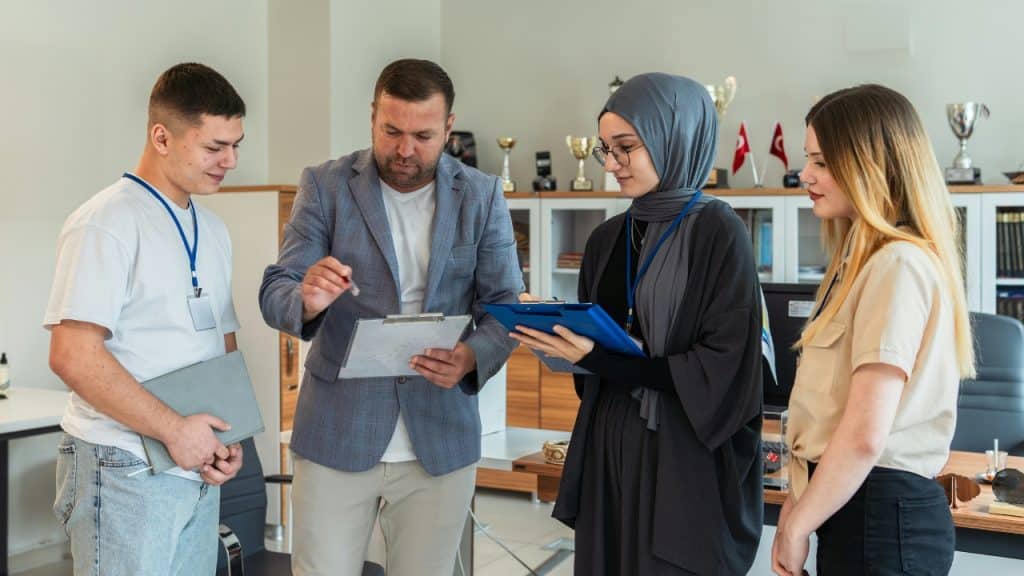 A man in a blazer holds a clipboard while three young adults stand around him.