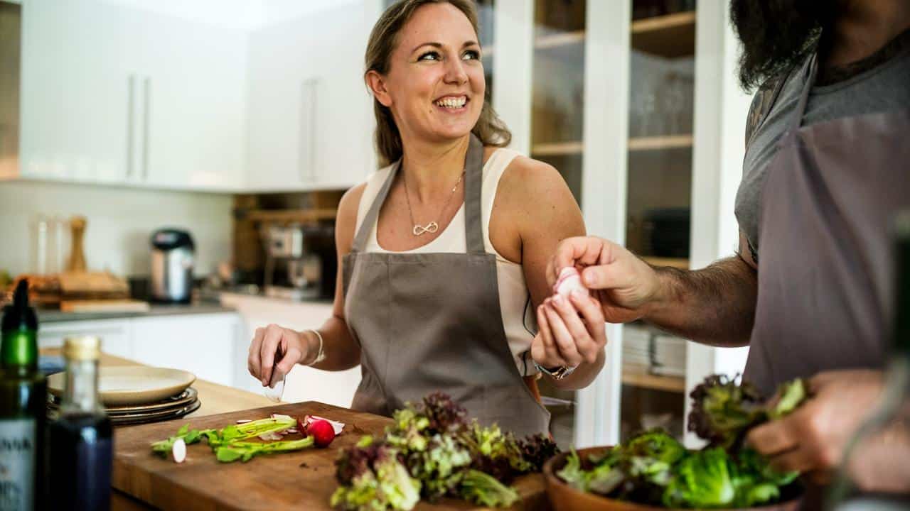 A smiling woman in an apron preparing vegetables with another person in a kitchen.