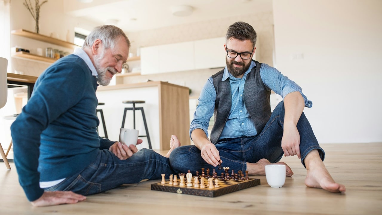 A cheerful adult hipster son and senior father sitting on the floor indoors at home playing chess.