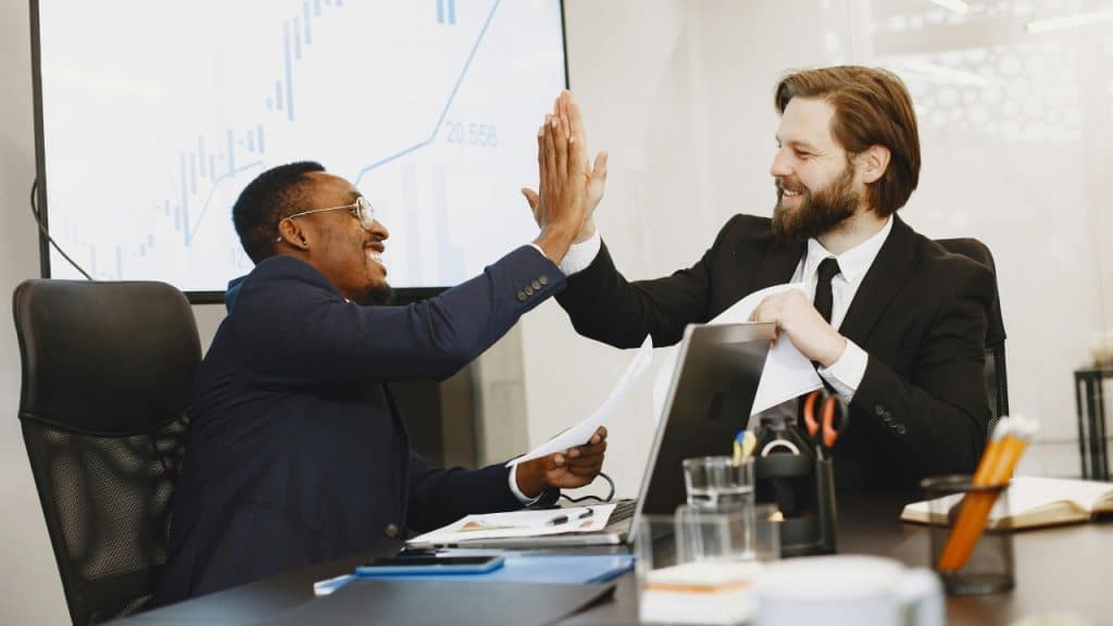 Two men in suits high-five each other across a desk in an office setting.