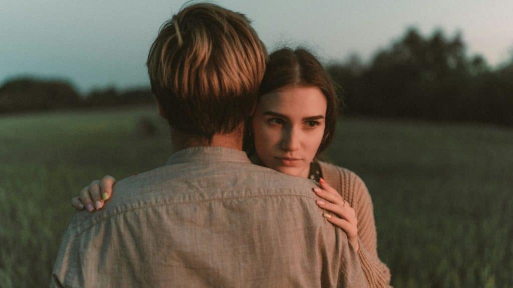 A woman rests her chin on a man's shoulder while looking into the distance with concern.