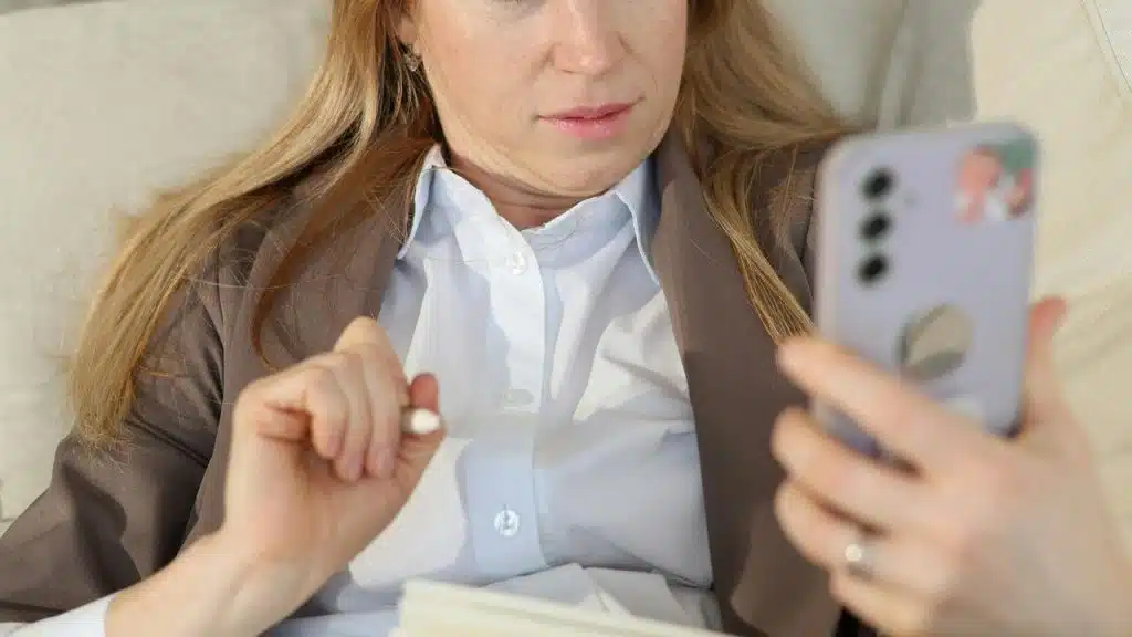 A woman looking at her phone while holding a pen and a notebook.