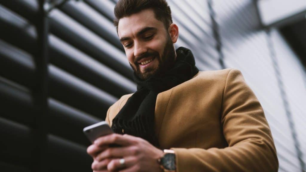 A man wearing a brown coat smiling while texting outdoors.