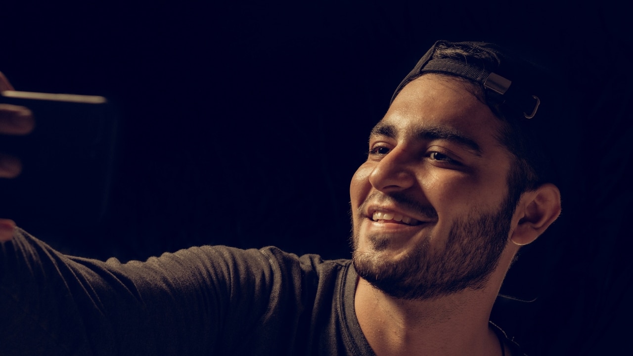 A man with a beard and baseball cap smiles while taking a selfie with a phone.