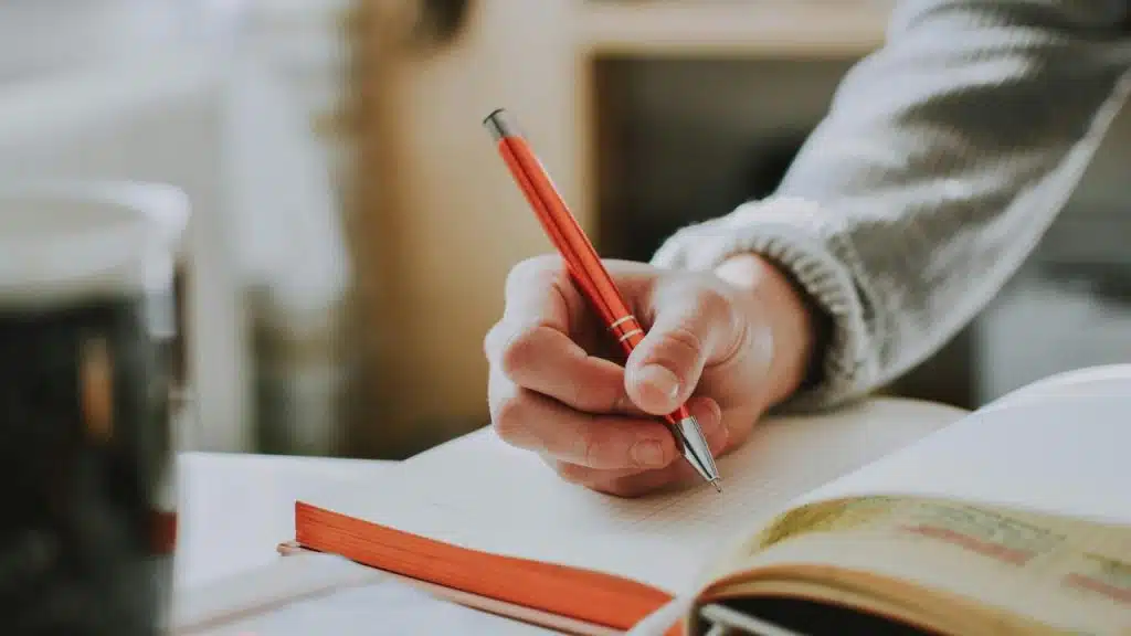 A person writes in a notebook with a red pen beside an open book.