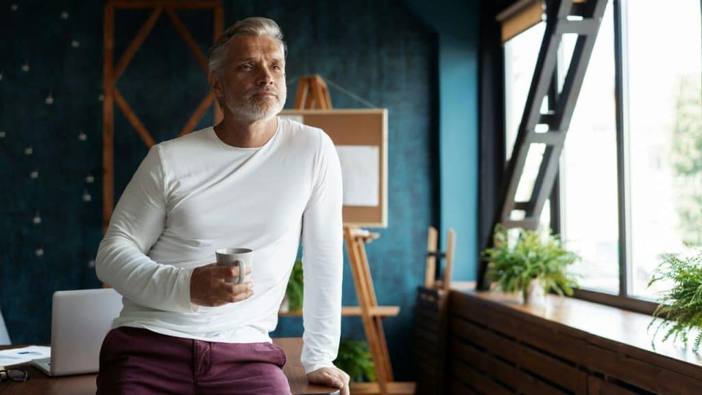 A gray-haired man in a white long-sleeve shirt leans on a desk holding a mug.