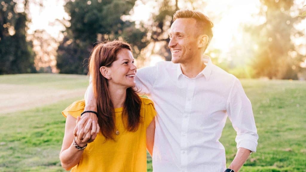 A smiling man and woman walk through a grassy field during a bright sunset.