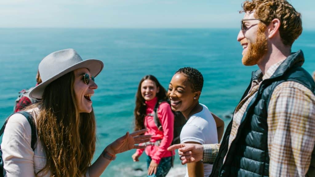 Four people with backpacks stand by the blue ocean and talk together on a sunny day.