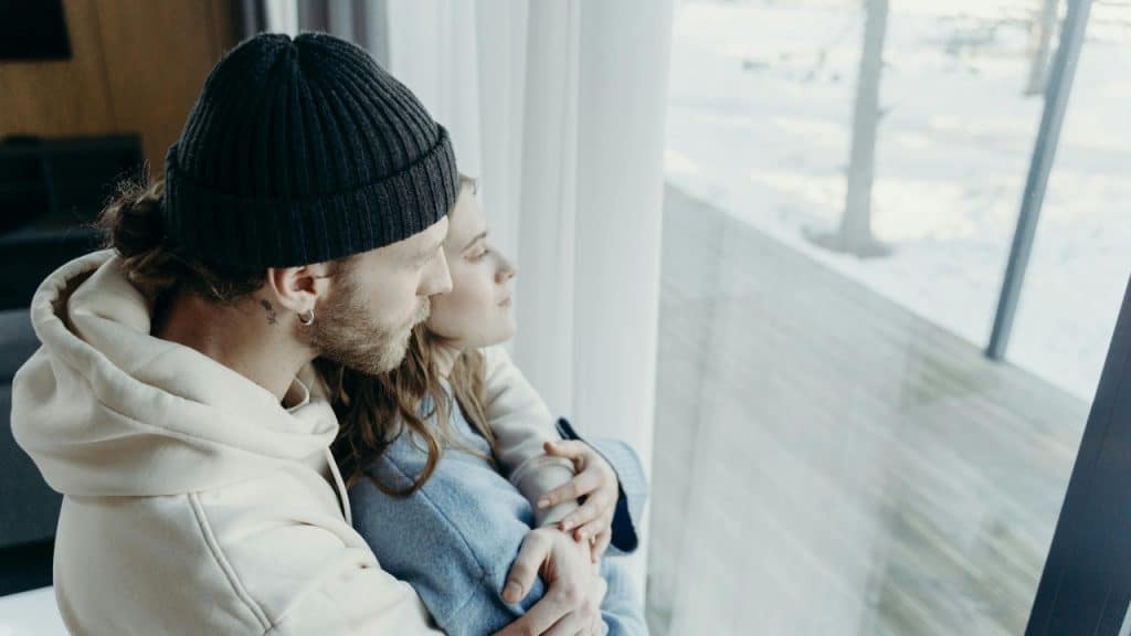 A man in a black beanie embraces a woman while they both look out a window.