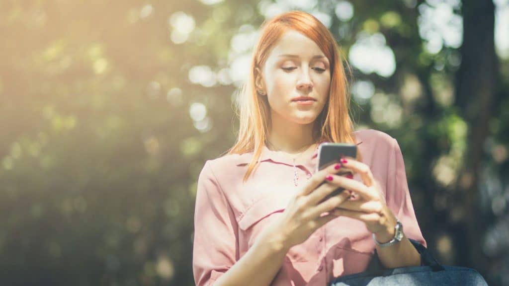 A woman with red hair looks down at her smartphone while standing outdoors.