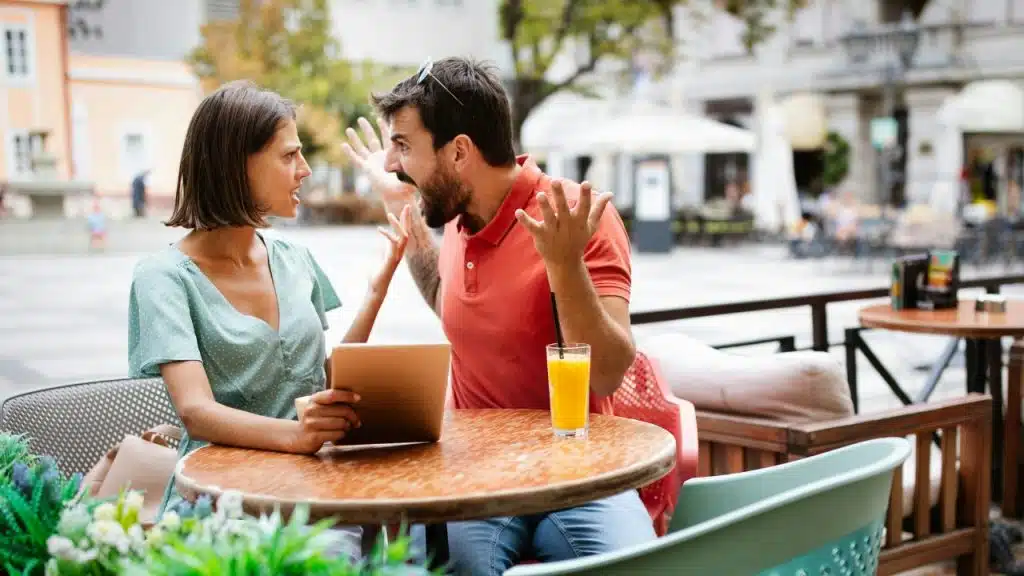 A man and woman sitting at an outdoor cafe table, gesturing with open hands while shouting.