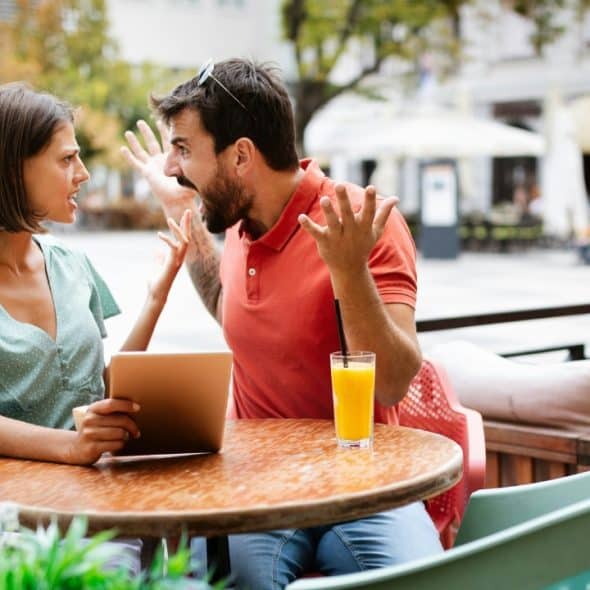 A man and woman sitting at an outdoor cafe table, gesturing with open hands while shouting.