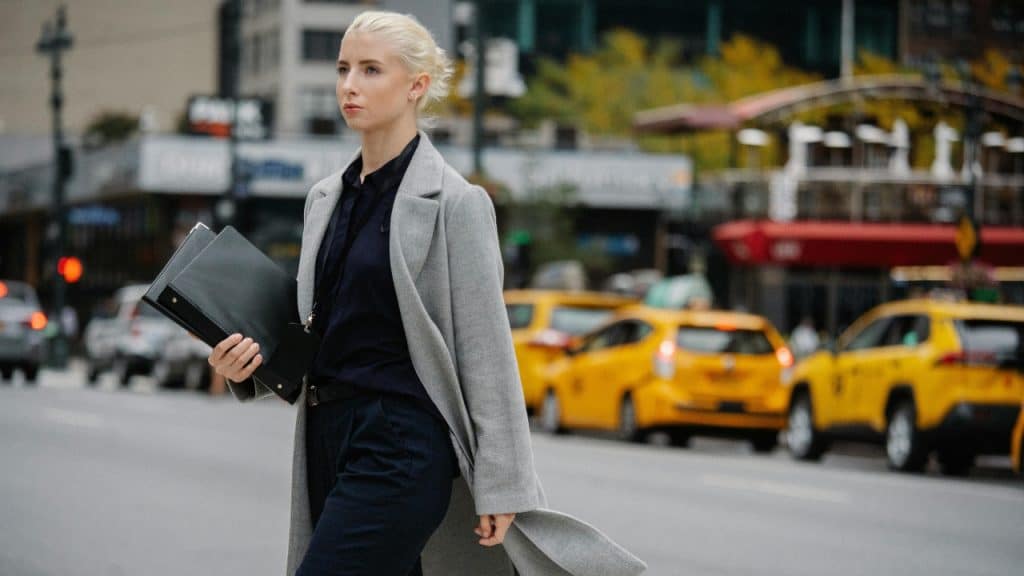 A woman in a grey coat walks across a city street with yellow taxis behind her.