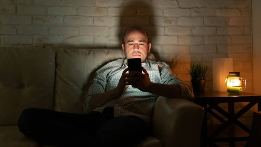 A man sits on a dark couch, his face illuminated by the screen of a smartphone.