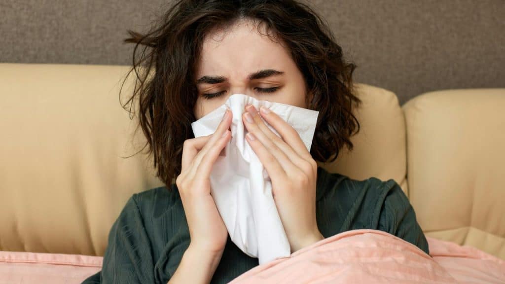 A woman sitting in bed blowing her nose with a tissue, appearing sick.