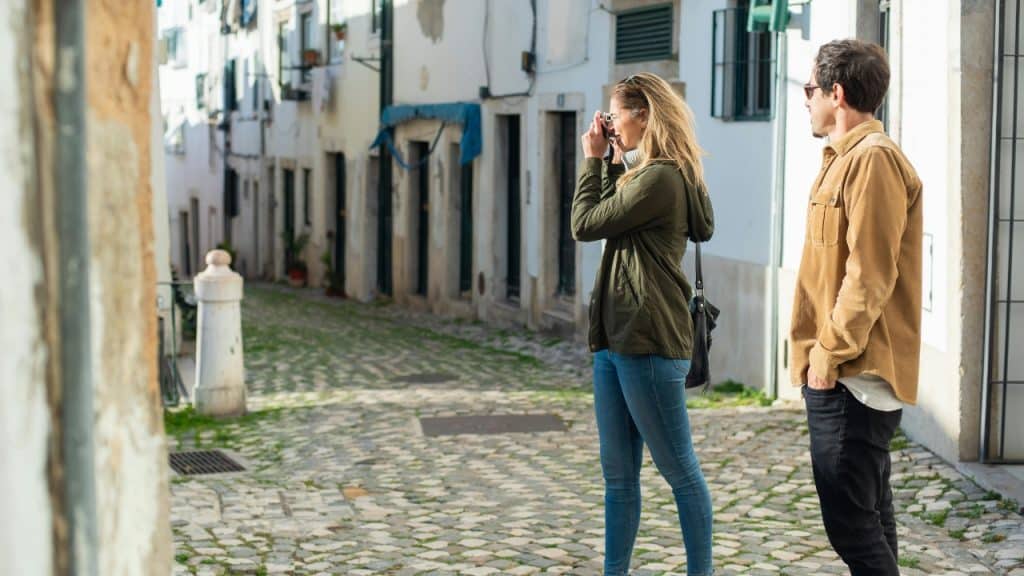 A woman in a green jacket takes a photo on a narrow cobblestone street alley.