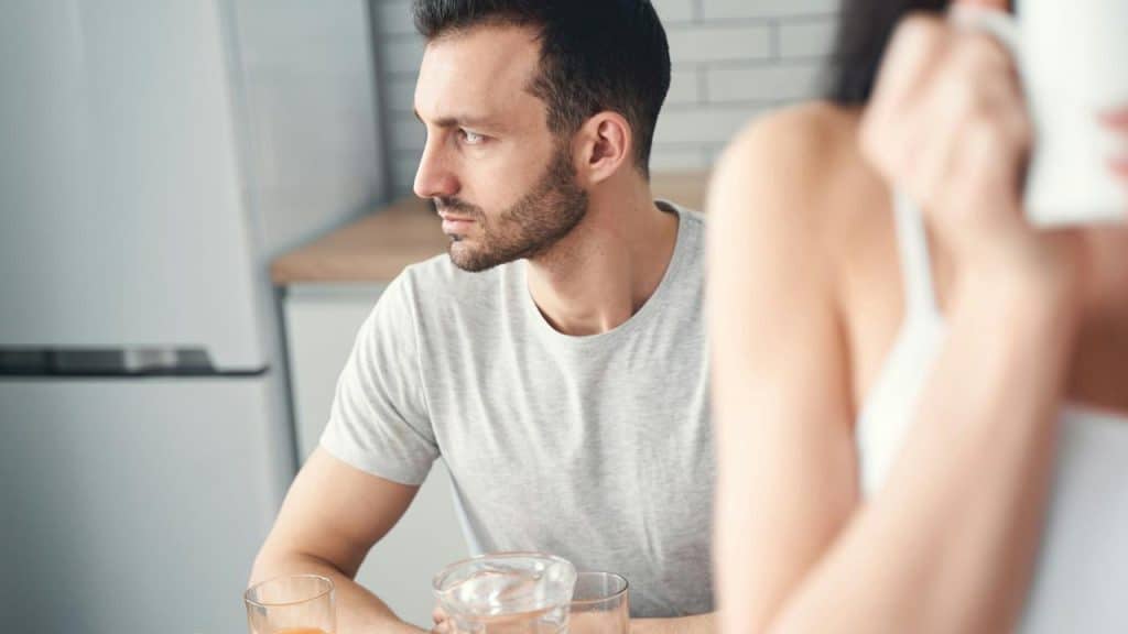 An upset bearded man looking to the side while a woman sips coffee.