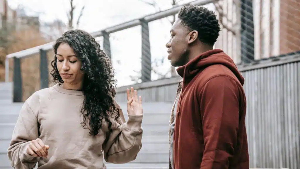 A woman with long curly hair is holding up her hand in a gesture of refusal or dismissal toward a man standing opposite her.