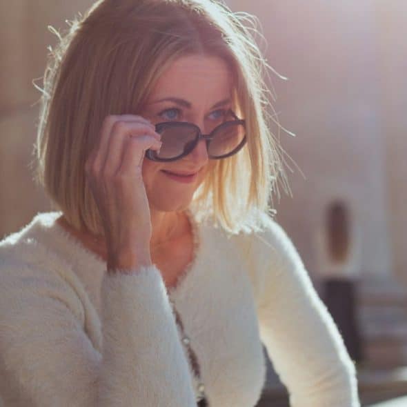 A woman with blonde hair and a white sweater lowers her sunglasses in the sunlight.