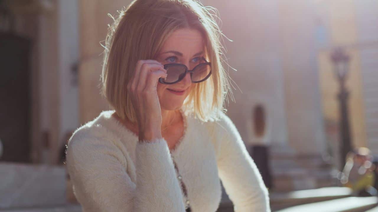 A woman with blonde hair and a white sweater lowers her sunglasses in the sunlight.