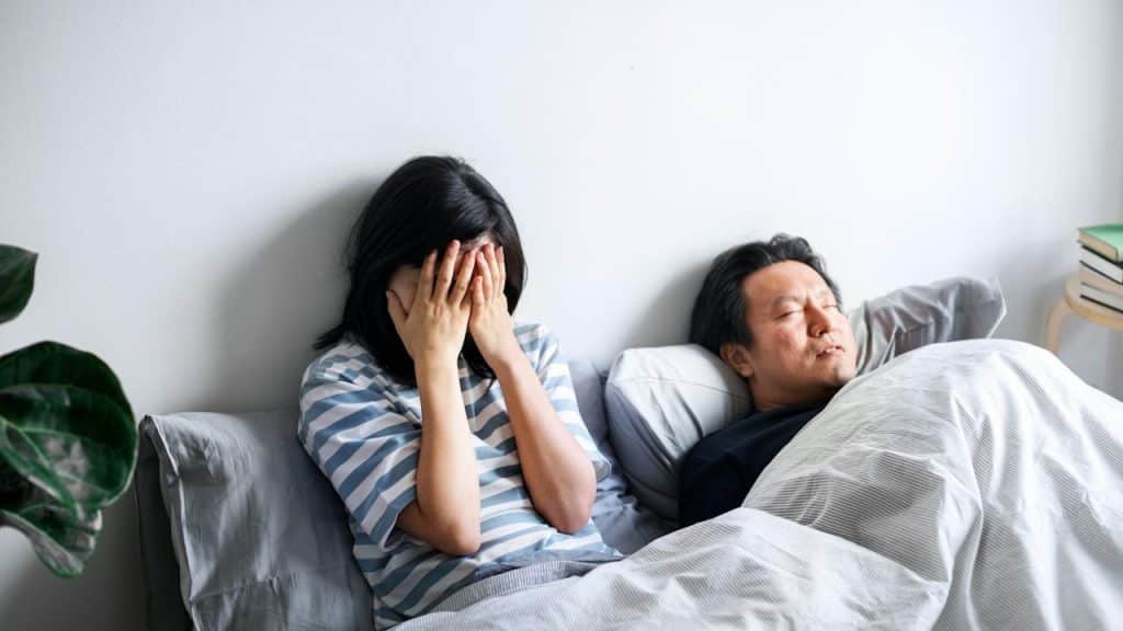 A stressed woman with hands on face siitin in bed with a man sleeping beside her.