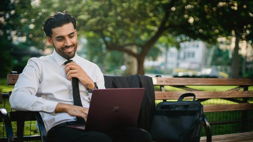 A man in a white shirt and tie sits on a bench using a laptop.