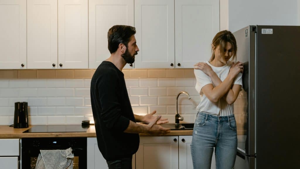 A man gestures with open hands toward a woman standing by a refrigerator in kitchen.
