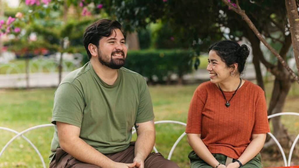 A man in a green t-shirt and a woman in an orange t-shirt sit on a bench together in a park, smiling and looking toward each other.
