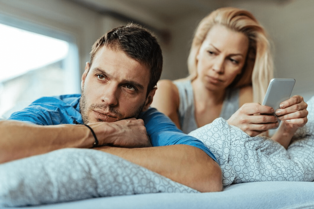 A displeased man is lying on the bed and ignoring his girlfriend