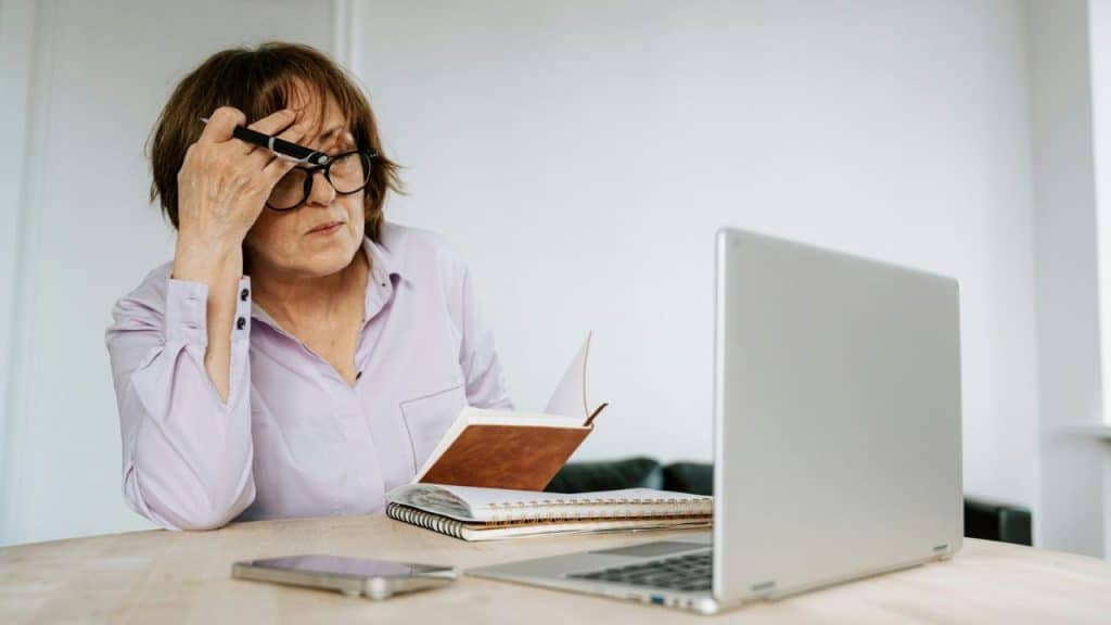 An older woman wearing glasses rests her head on her hand while looking at a laptop.