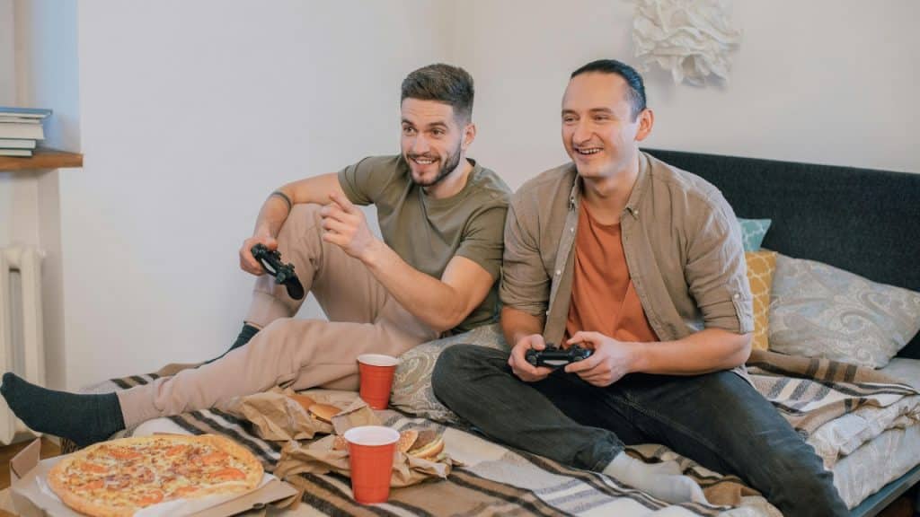 Two men sit on a bed holding controllers near pizza, burgers, and red plastic cups.