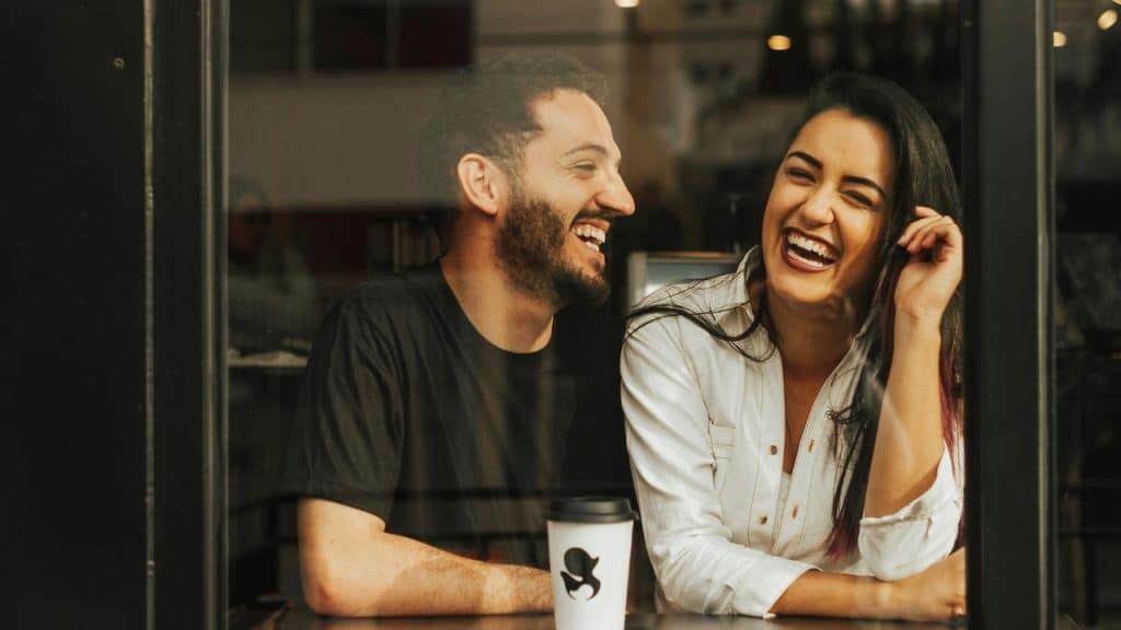 A man and woman laugh together while sitting at a table with coffee.