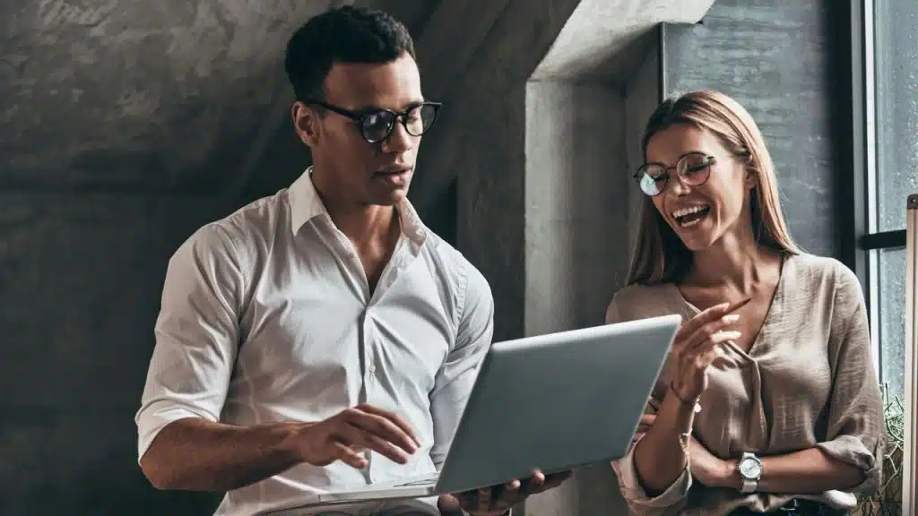 A woman laughs and points toward a man who is looking at a laptop screen.