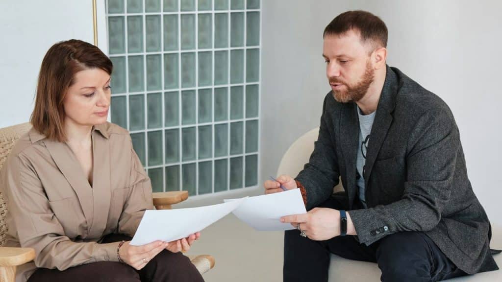 A man and woman sit together indoors, both looking down at several white papers.