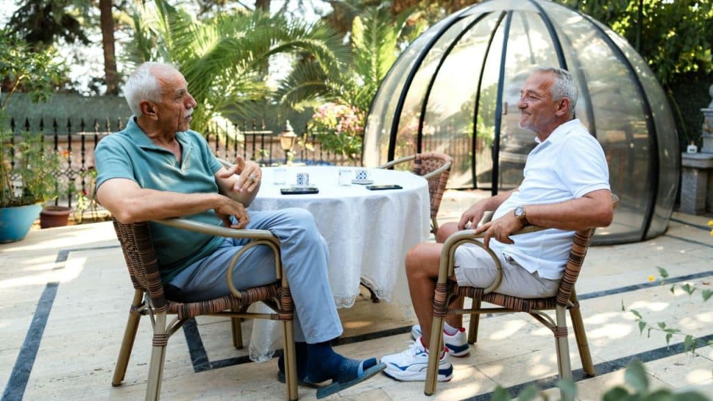 Two older men sit in chairs at a white table outdoors, talking near a glass dome.