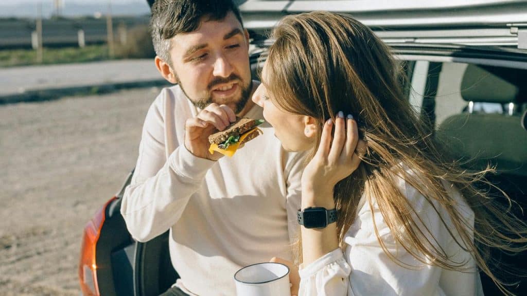 A couple sitting in a car trunk, with the man feeding the woman.