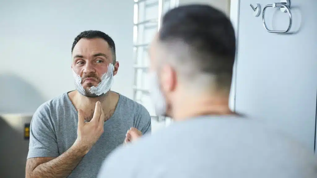A man with shaving cream on his face looks into a mirror while grooming his chin.