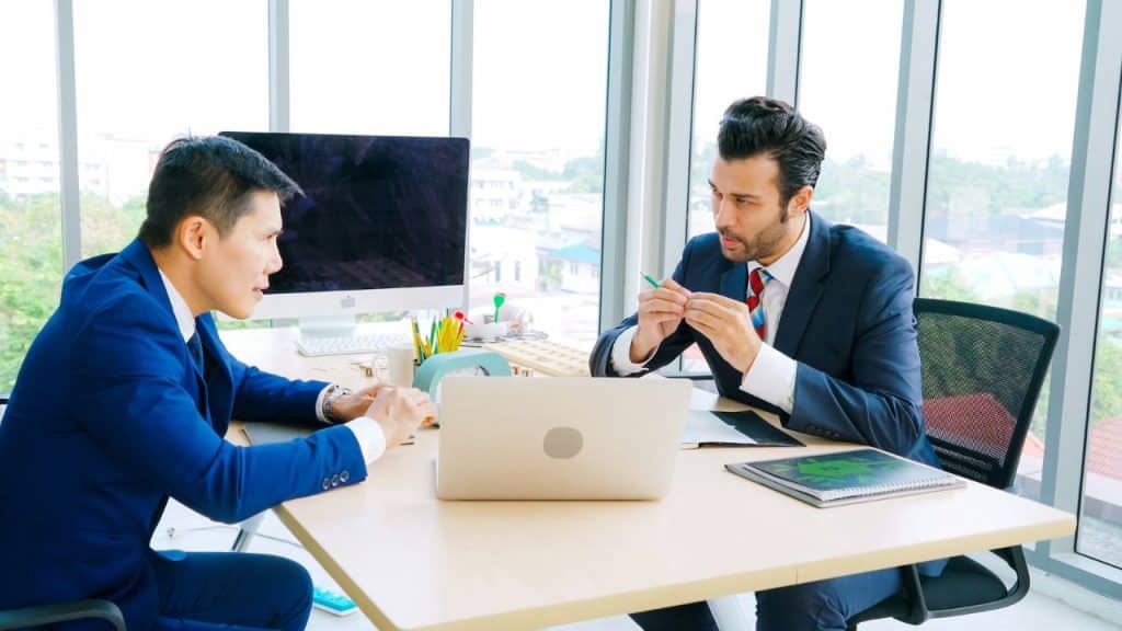 Two men in suits sitting at a desk and discussing work in an office.