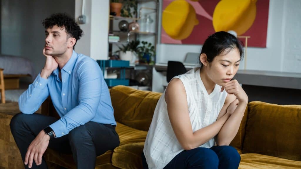 A man and woman sit on a yellow sofa, each looking in opposite directions.